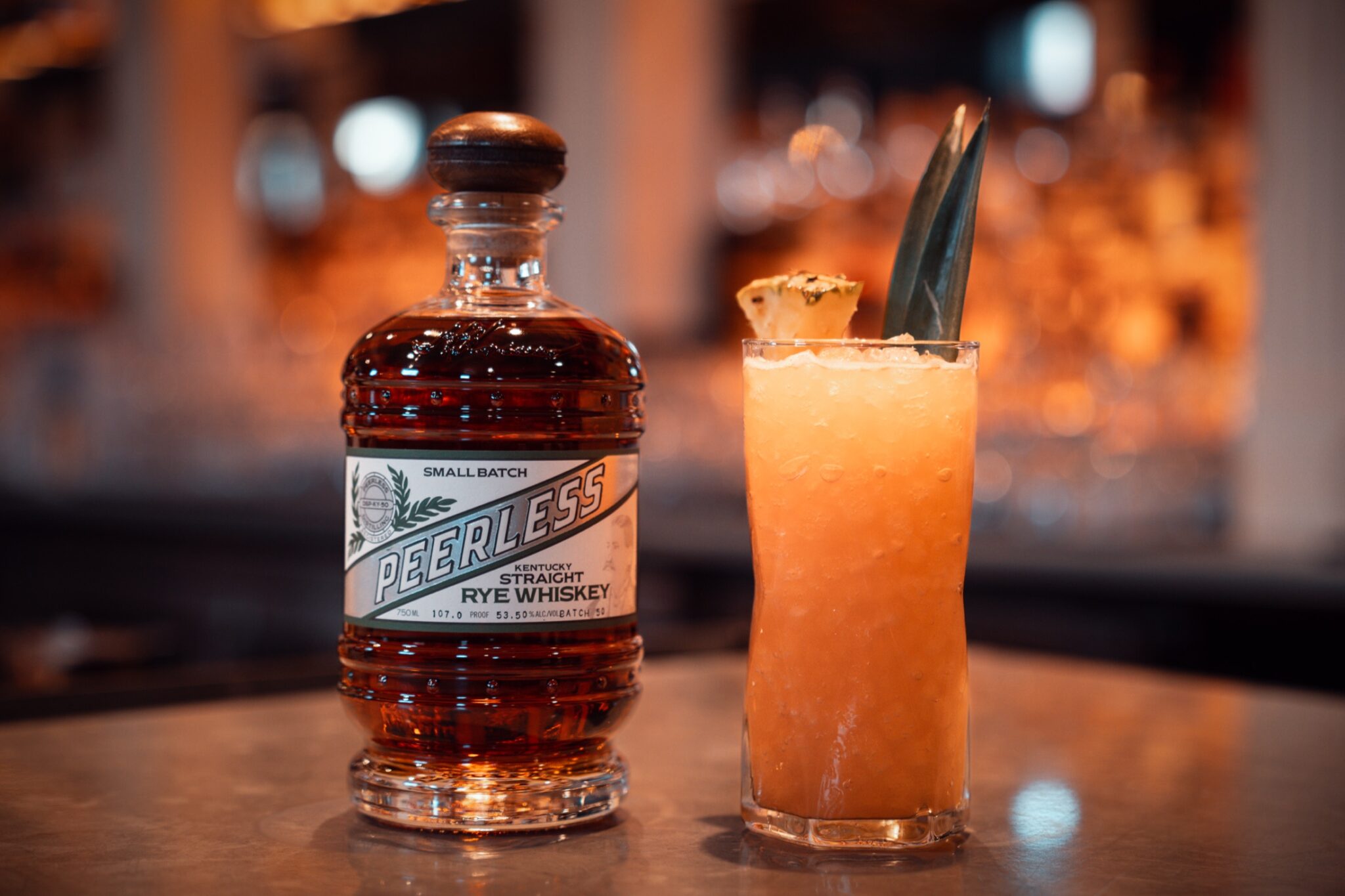 Peerless small batch rye bottle beside a pineapple-garnished cocktail at a warm bar